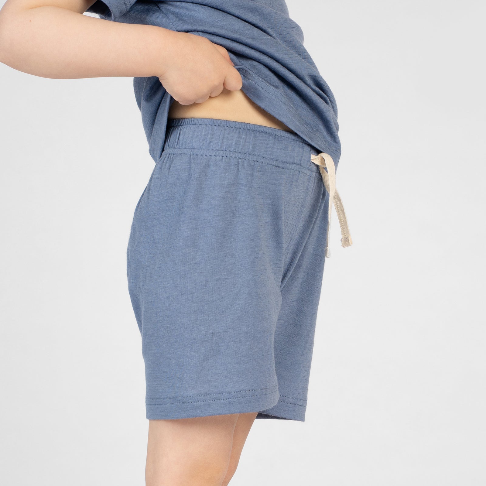 A close-up side profile view of a young child's torso and upper legs. The child is wearing a mustard yellow merino short-sleeve t-shirt and matching mustard yellow merino shorts. The child's hand is slightly lifting the hem of the t-shirt to show the elastic waistband and drawstring of the shorts.