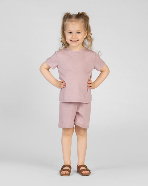 A smiling young girl with blonde pigtails stands facing the camera with her hands on her hips against a light gray background. She is wearing a dusty pink merino short-sleeve t-shirt, matching dusty pink merino shorts, and brown strappy sandals.