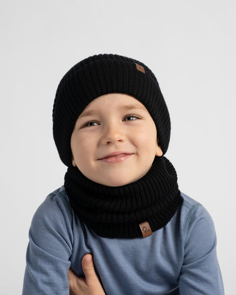 A chest-up portrait of a child looking slightly upward with a smile, wearing a slate blue shirt, black neck gaiter, and black knit beanie.