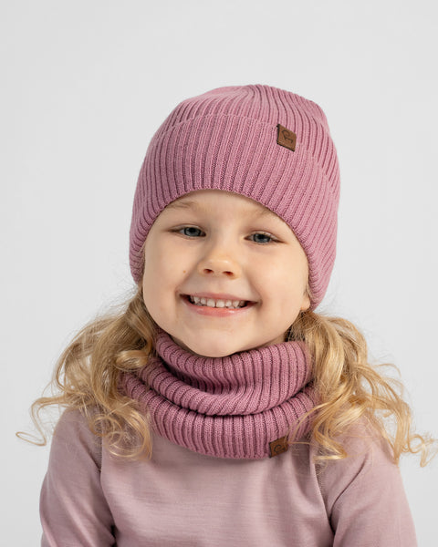 A smiling young girl with wavy hair wearing a dusty rose-pink knit beanie and matching neck gaiter.