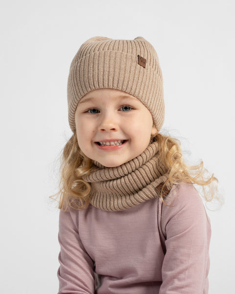 A chest-up portrait of a smiling young girl wearing a dusty rose top, matching beige knit neck warmer, and a light beige beanie.