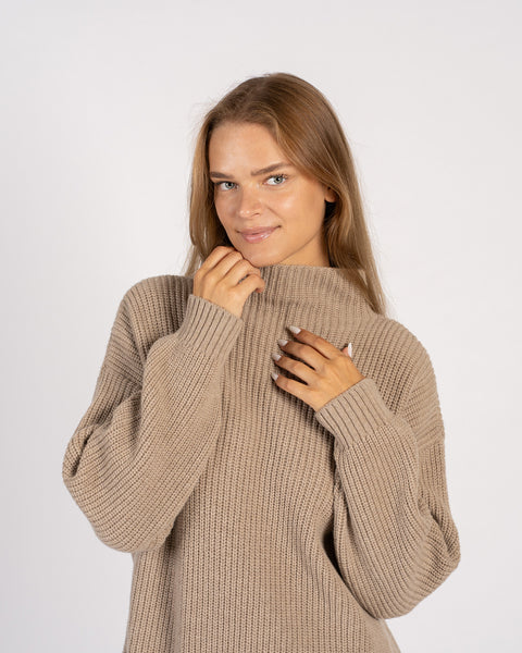 Close-up of a woman gently holding the high neck of a beige knit sweater, showcasing the cozy ribbed texture.