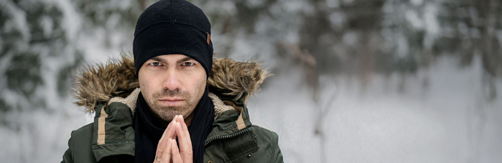 Man wearing a warm jacket with fur hood and black beanie standing outdoors in a snowy forest with hands clasped near face