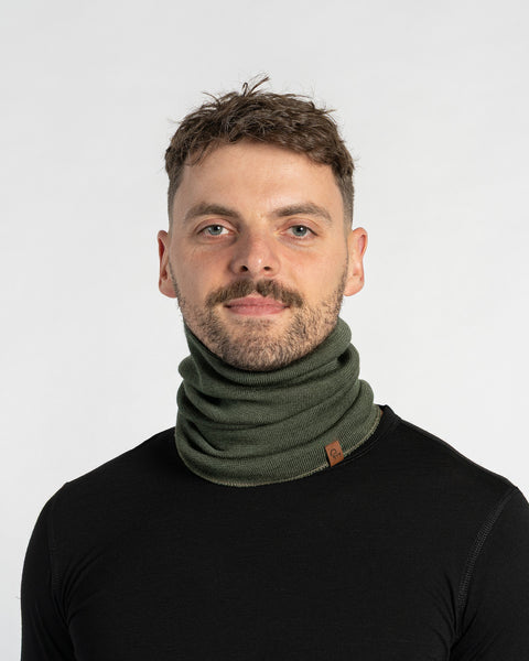 A man with short dark hair, a mustache, and a beard looks at the camera while modeling a dark green knitted neck gaiter. He is wearing a black shirt, and the gaiter is bunched around his neck against a plain white background.