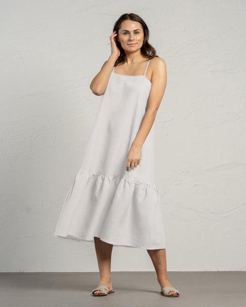 A woman with long, dark hair stands in a full-length shot, facing slightly to her left, wearing a menique pure white linen dress with thin straps and a ruffled hem. Her right hand is gently touching her hair near her ear, while her left arm hangs loosely at her side. She is wearing light-colored, open-toe slip-on sandals. The background is a textured light grey wall and a darker grey floor. The lighting is soft and even, minimizing shadows.