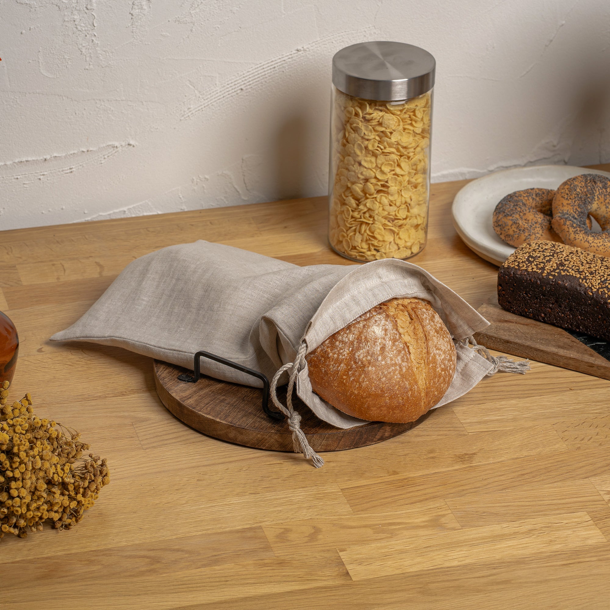 A bagel sprinkled with poppy seeds rests inside a natural color linen drawstring bag, which is open and draped over a round wooden board with a dark metal handle. The board sits on a light-colored wooden countertop. In the background, partially visible, are a glass jar filled with pasta, a round loaf of bread on a white plate, and a dark loaf on a rectangular wooden cutting board. Some dried herbs are scattered in the bottom left corner of the frame.