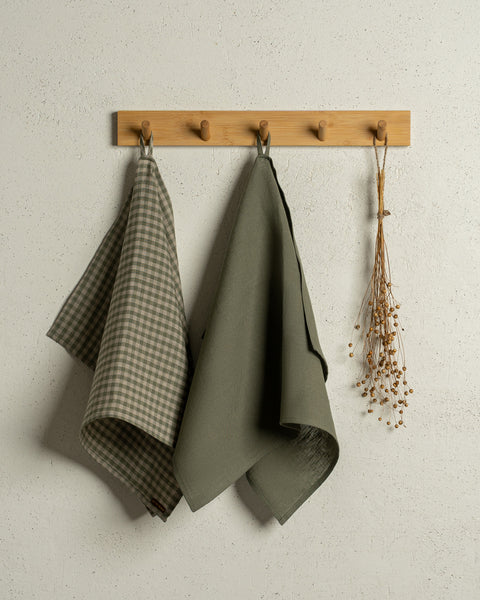 Two menique linen kitchen dish towels hanging on a wooden rack on a wall. The colors of the towels are stone green and green check. 