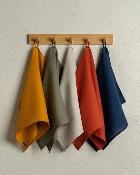 The menique linen kitchen dish towels in the colors of spicy yellow, stone green, natural, cinnamon red, and storm blue hanging on a wooden rack on a wall.
