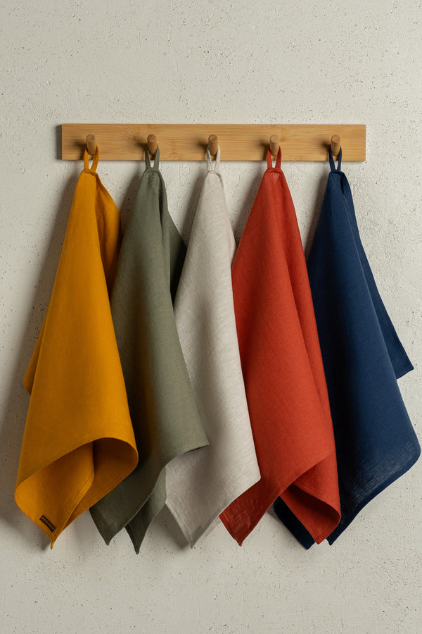 The menique linen kitchen dish towels in the colors of spicy yellow, stone green, natural, cinnamon red, and storm blue hanging on a wooden rack on a wall.