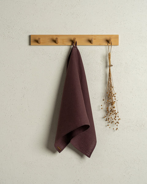 The menique linen kitchen dish towel in the color of shadow purple hanging on a wooden rack on a wall.
