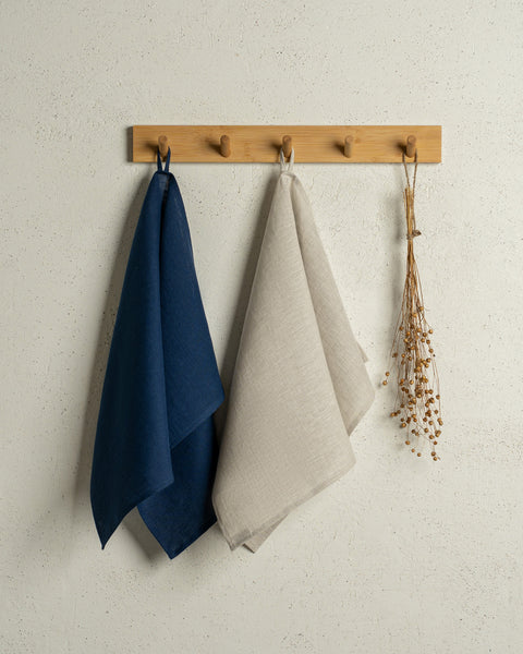Two menique linen kitchen dish towels hanging on a wooden rack on a wall. The colors of the towels are storm blue and natural. 