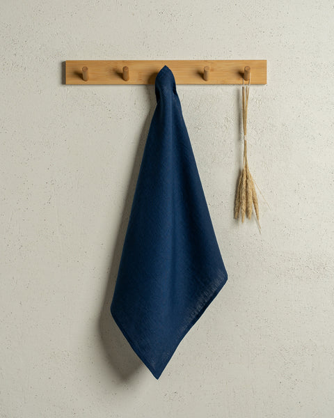 The menique square linen dish towel in color storm blue hanging on a wooden rack on a wall.