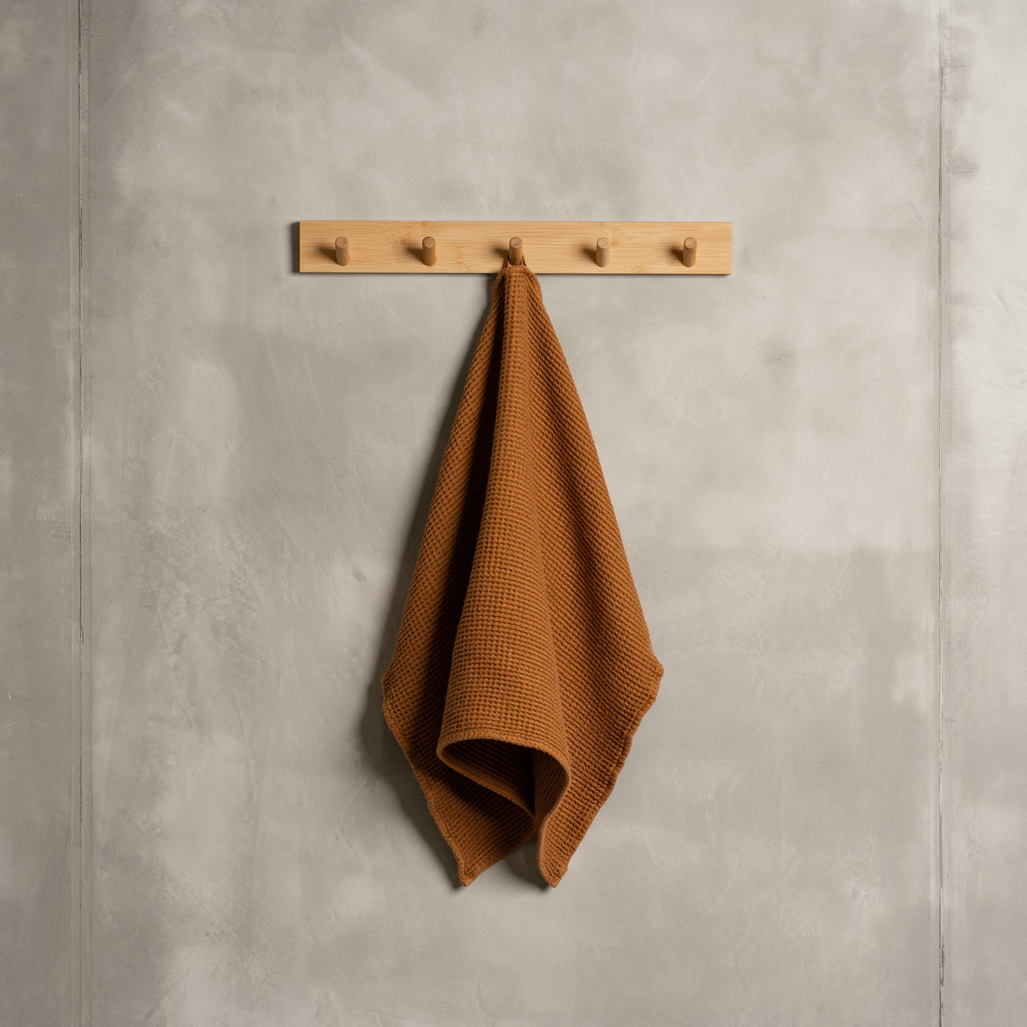 A gray wall with a wooden hanger. In the middle of it hangs a waffle hand washcloth in stone green color.