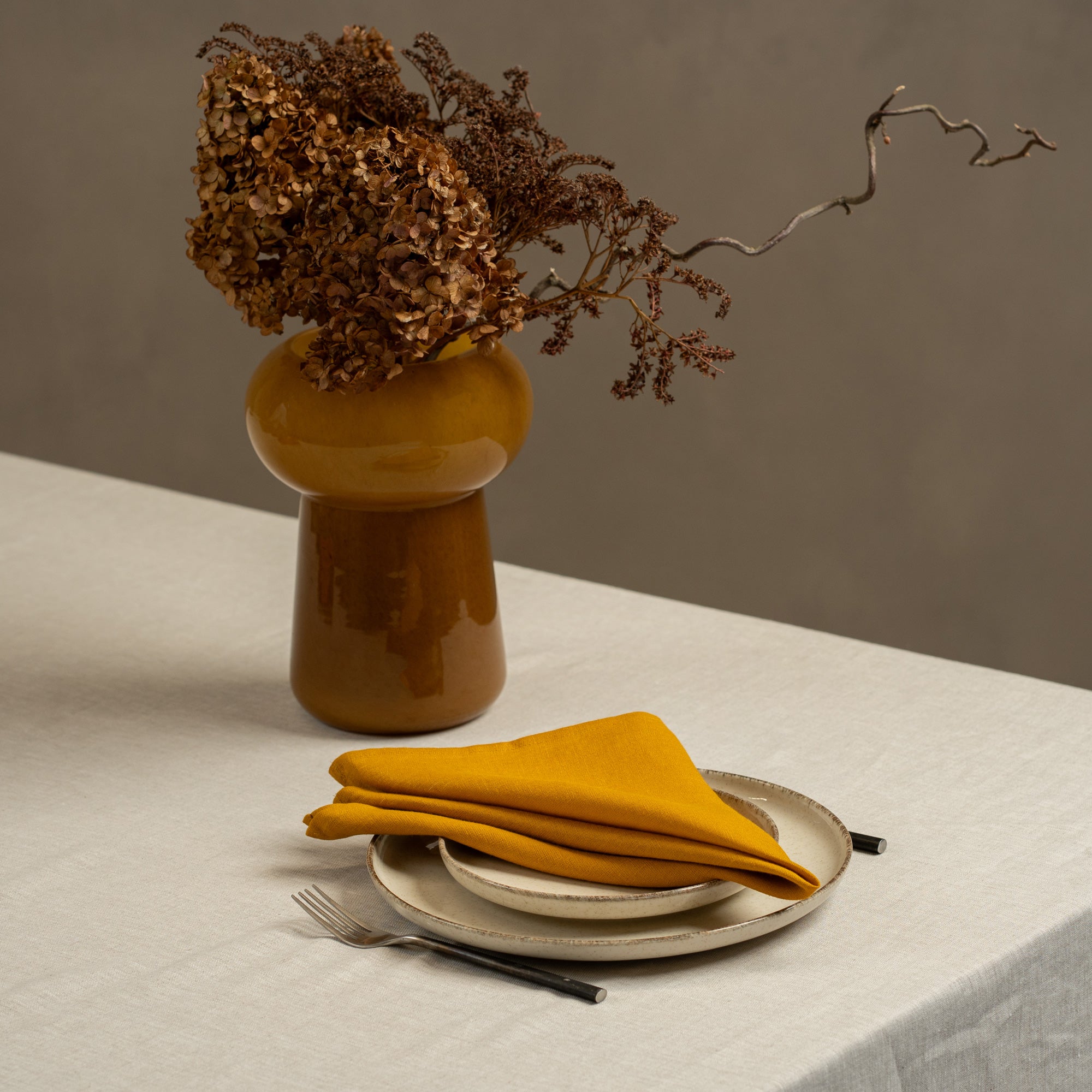 A tabletop covered with a linen tablecloth, with a vase and a single plate set on top of it. By the sides of the plates there are cutlery items, and there is a linen napkin in dark green color on top.