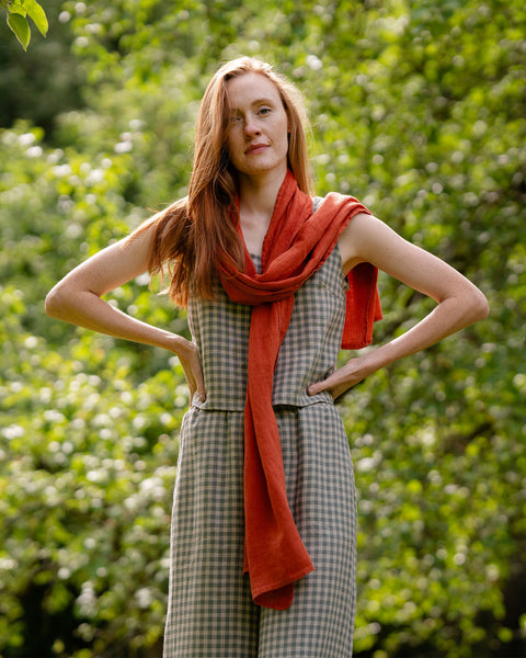 A woman wearing a checkered outfit and an orange scarf poses with their hands on their hips against a backdrop of green leaves.
