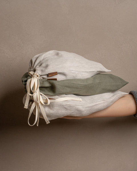 A hand wearing a gray knitted sleeve holds a stack of three closed linen drawstring bags in stone green and natural colors against a warm brown background.