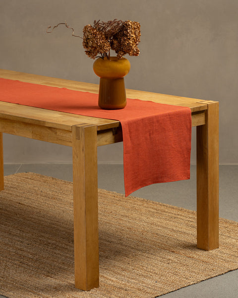The cinnamon red linen table runner on top of a wooden table, captured in a close up picture from the corner of the table. 