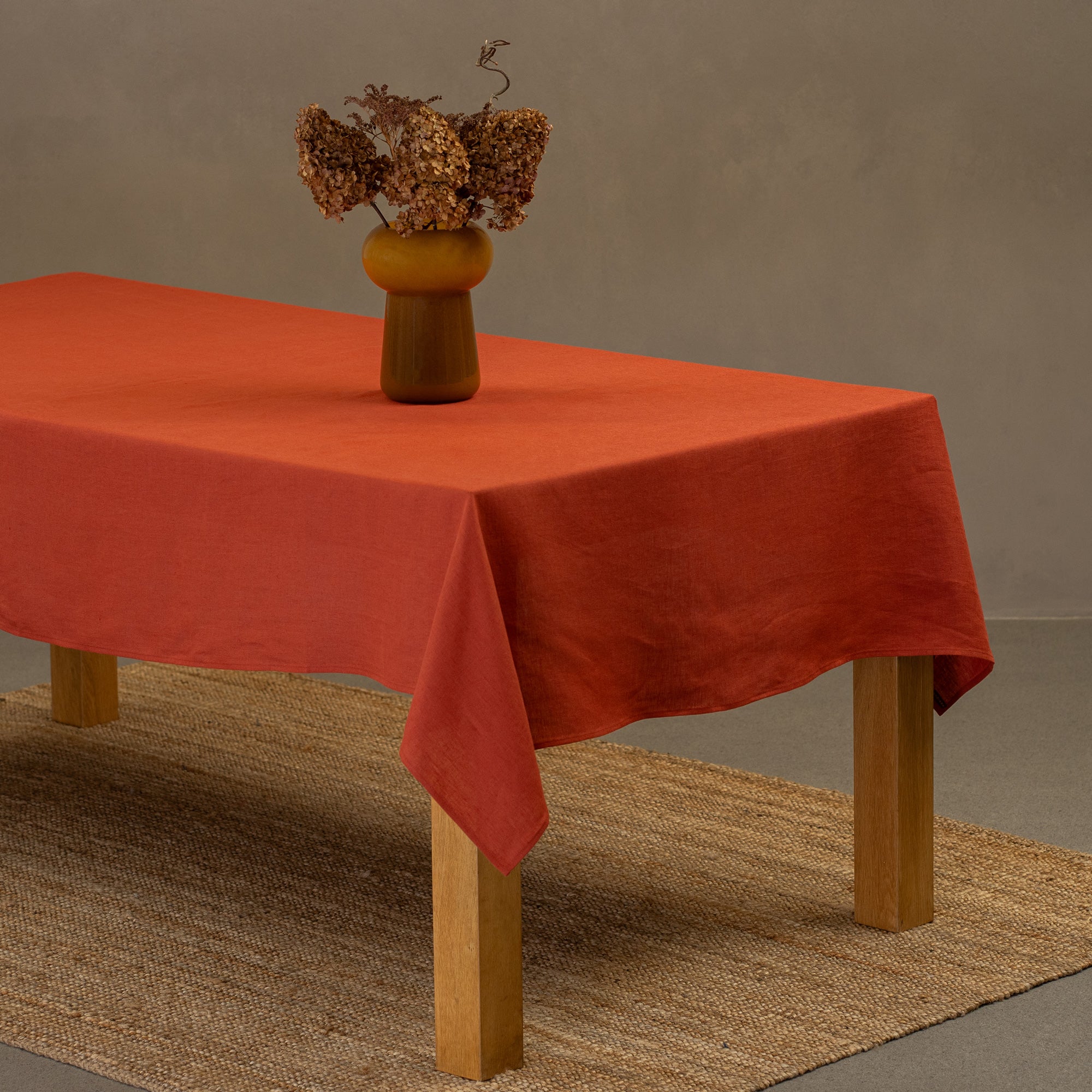 The menique stone green linen tablecloth on a wooden table. On top of the tablecloth is a vase made of natural straws and a clay cup. Under the table is a carpet.