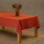 The menique stone green linen tablecloth on a wooden table. On top of the tablecloth is a vase made of natural straws and a clay cup. Under the table is a carpet.