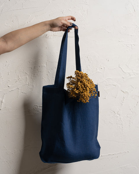 A fair-skinned arm and hand hold up a storm blue reusable linen tote bag against a textured off-white wall. Dried yellow flowers are visible inside the bag.