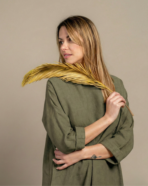Woman wearing stone green linen dress, holding dried palm leaf, minimalist studio background.