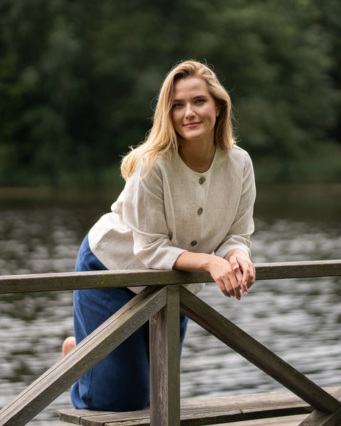 A smiling blonde woman leans comfortably against a wooden railing beside a calm body of water. She is casually dressed in a light cream sweater and dark blue denim.