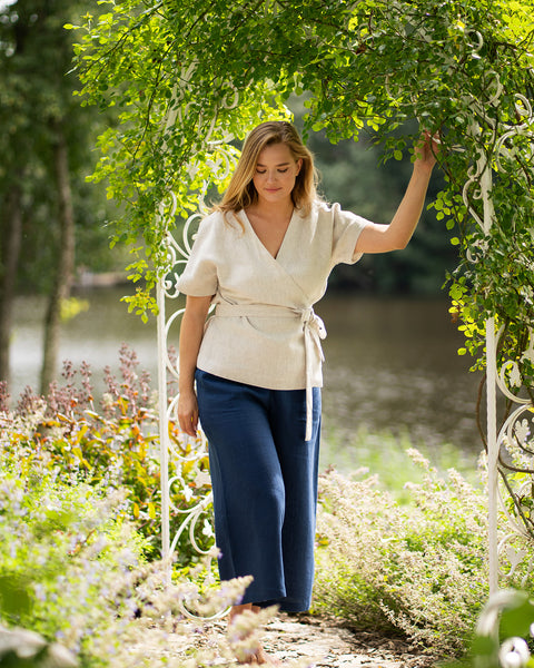 A barefoot woman walks thoughtfully through a white metal garden archway surrounded by lush greenery and flowers. She is dressed in a light beige, short-sleeved linen wrap top and wide-leg storm blue pants.