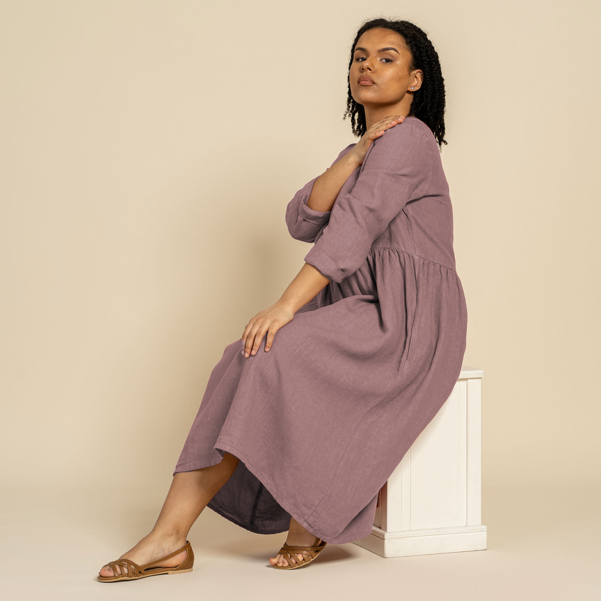 A woman with dark curly hair stands looking at the camera, wearing a menique linen long-sleeved dress in the color faded rose. Her hands are in the side pockets of the dress. She is positioned against a solid light brown background.