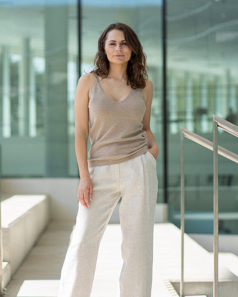 A woman with shoulder-length wavy brown hair stands looking directly at the camera, with one hand in her pocket and the other relaxed at her side. She is wearing a merino wool and cashmere tank top and linen menique wide-legged trousers, and is positioned on light-colored stairs with silver handrails and a modern building with large windows in the background.