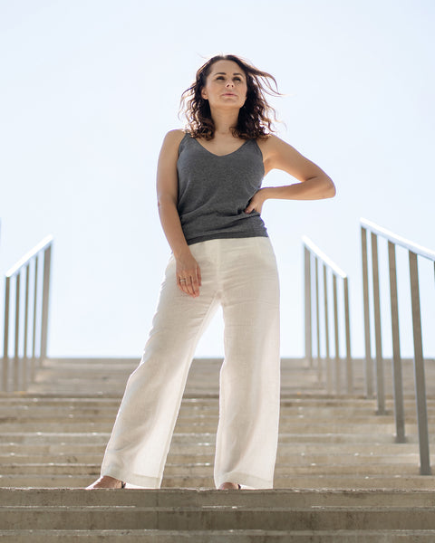 A woman with wavy brown hair stands confidently on outdoor concrete steps, looking slightly upwards with one hand on her hip. She is wearing a dark gray tank top and menique linen white trousers, with metal railings on either side of the stairs leading up to a bright sky.