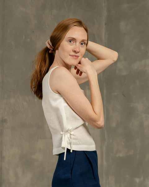 A studio portrait of a young woman with reddish-brown hair pulled back in a low ponytail, looking towards the viewer with a slight smile. She is wearing a white sleeveless linen top with side ties at the hem. Her left arm is bent with her hand near her chin, and her right arm is bent with her hand near the back of her head. The background is a textured grey wall.