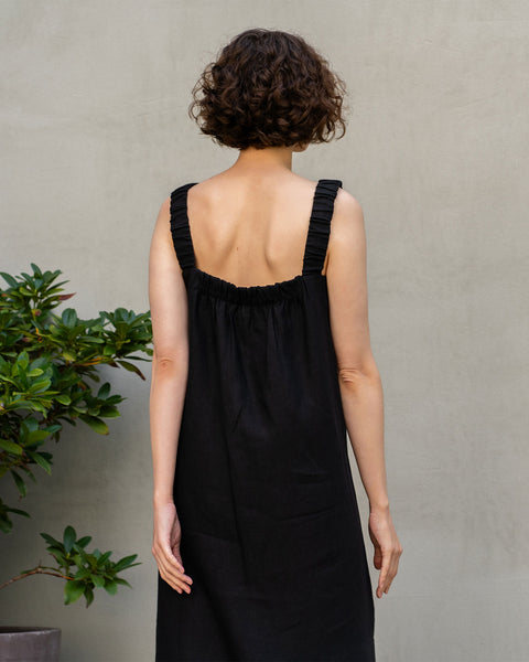 A fair-skinned woman with short, dark curly hair stands facing away from the viewer. She is wearing a sleeveless pure black linen maxi dress with gathered details at the back. To her left is a leafy green plant in a pot, and she is standing against a plain, light-colored wall.