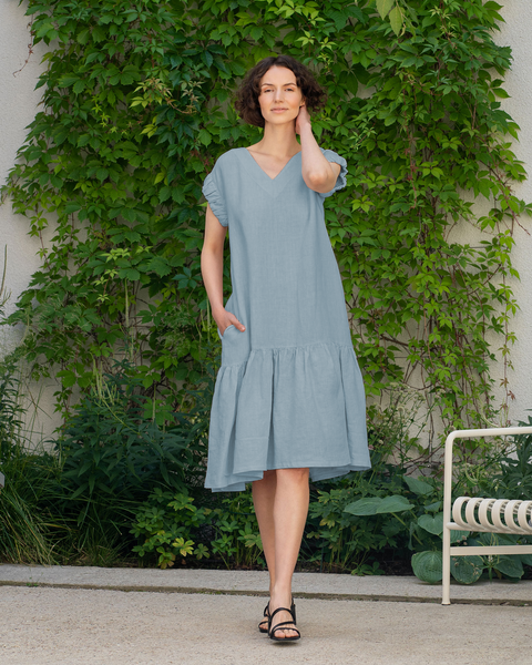 A woman with short, dark curly hair stands outdoors, looking to the right. She is wearing menique cloudy blue, linen dress. Her left hand is in the dress pocket, and her right hand is near her neck. She is standing on a light-colored paved surface in front of a wall covered with green ivy. A white metal chair is partially visible on the right side of the frame, surrounded by various green plants. 