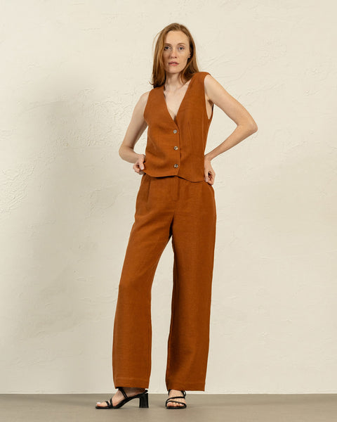 A person with shoulder-length brown hair stands facing forward, modeling an almond brown linen vest and menique wide-legged trousers. They have their hands resting on their hips and are wearing black open-toe sandals with a low heel. The background is a textured light beige wall and a light gray floor.