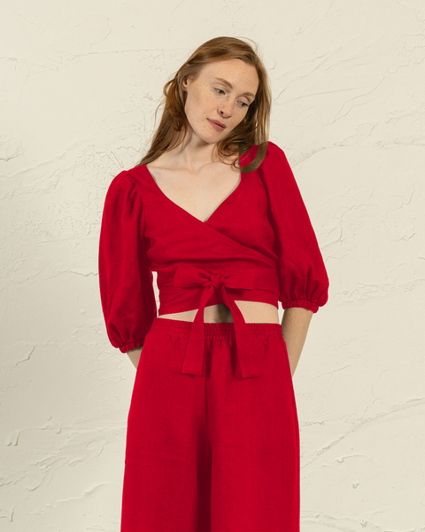A light-skinned person with long, reddish-brown hair poses against a textured off-white background, wearing menique pure red wrap top with a deep V-neck that wraps and ties at the waist, featuring three-quarter length puffed sleeves. The person is looking down and slightly to the right, with their hands gently resting at their sides and partially obscured by the trousers. 