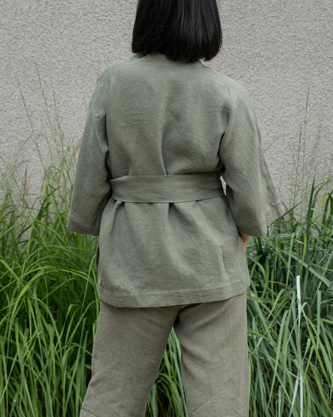 Woman with short dark hair wearing a stone green linen jacket and stone green menique pants, viewed from the back.