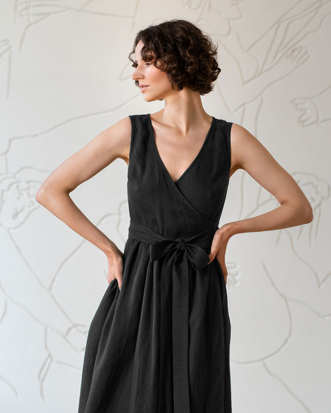 A woman with short brown hair is wearing menique sleeveless linen pure black dress with a V-neck. A matching fabric belt is tied loosely at her waist. Her hands is gently placed on her hips. 