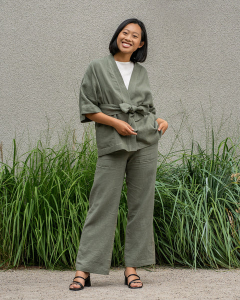 A person stands outdoors, wearing a stone green colored linen kimono-style top and matching wide-leg pants. Their right hand is in the pocket of their pants. To their left, there are tall green plants.