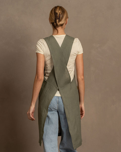 A woman with her back to the viewer shows the cross-back design of a stone green linen cooking apron, wearing a white t-shirt and light blue jeans.