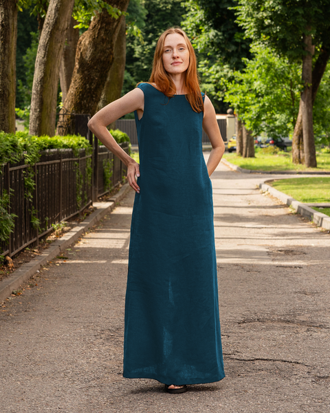 A woman with long reddish hair in a menique linen sleeveless dress stands on a path in a park.