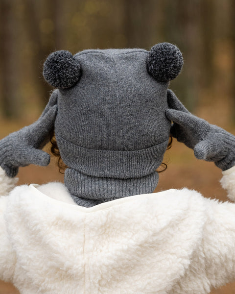 The back of a child&#39;s head and shoulders are shown, with the child wearing a dark grey knit hat with two dark grey pom-poms on top. They are also wearing a grey turtleneck and a cream-colored sherpa jacket, with dark grey mittens on their hands, against a blurred forest background.