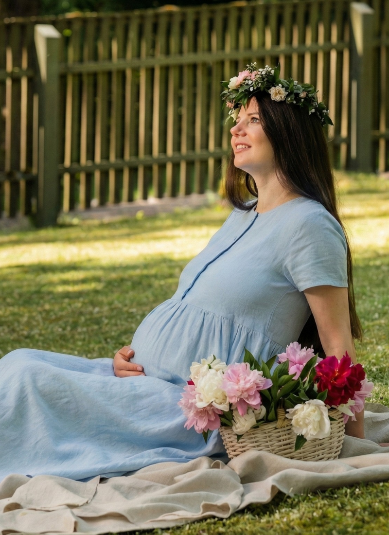 A pregnant woman is sitting on a blanket in a green garden, wearing a light blue linen maternity dress and a delicate flower crown. She gently rests her hand on her baby bump while looking upward, with a basket of fresh flowers beside her, creating a serene and joyful summer scene.