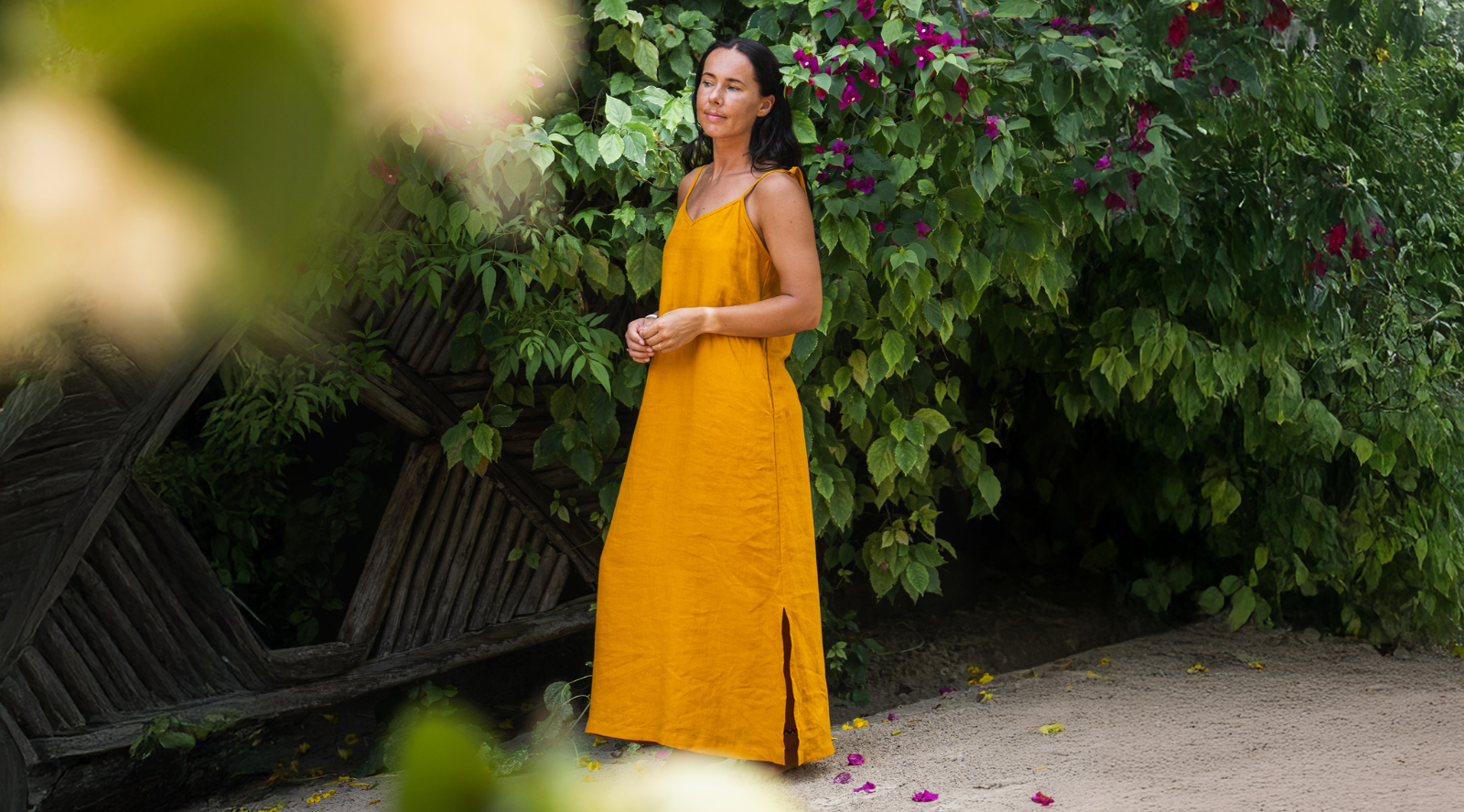 Woman wearing a mustard yellow linen maxi dress standing in a lush garden with green foliage and flowers.