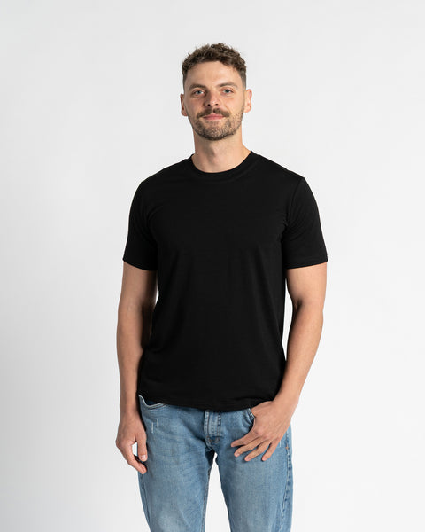 A man with short, dark brown hair and a neatly trimmed beard stands centered against a plain white background, directly facing the viewer. He wears a black crew neck t-shirt with short sleeves from menique brand, showcasing the garment&#39;s fit and simplicity. The t-shirt is made of merino wool fabric. He is also wearing light blue denim jeans with a straight leg cut and a visible front pocket, with one hand casually tucked into the pocket. 