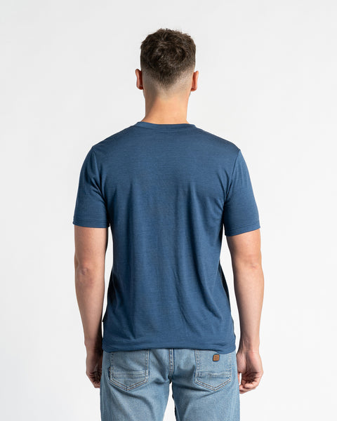 Back view of a man wearing a menique merino wool t-shirt and light blue jeans, standing against a white background. The denim color t-shirt is the primary focus, showcasing its fit and design from the rear. The man is positioned centrally, displaying the back of the garment clearly.