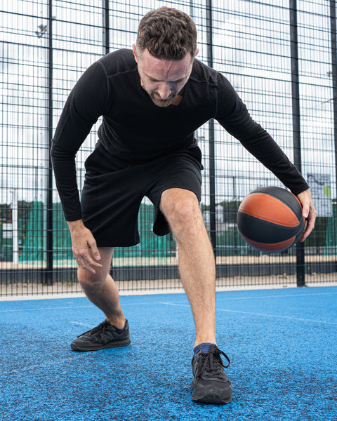 A man on a blue sports court in a low stance, dribbling an orange and black basketball with his right hand. He is wearing a black long-sleeved shirt, black shorts, and dark athletic shoes, with a tall wire fence in the background.