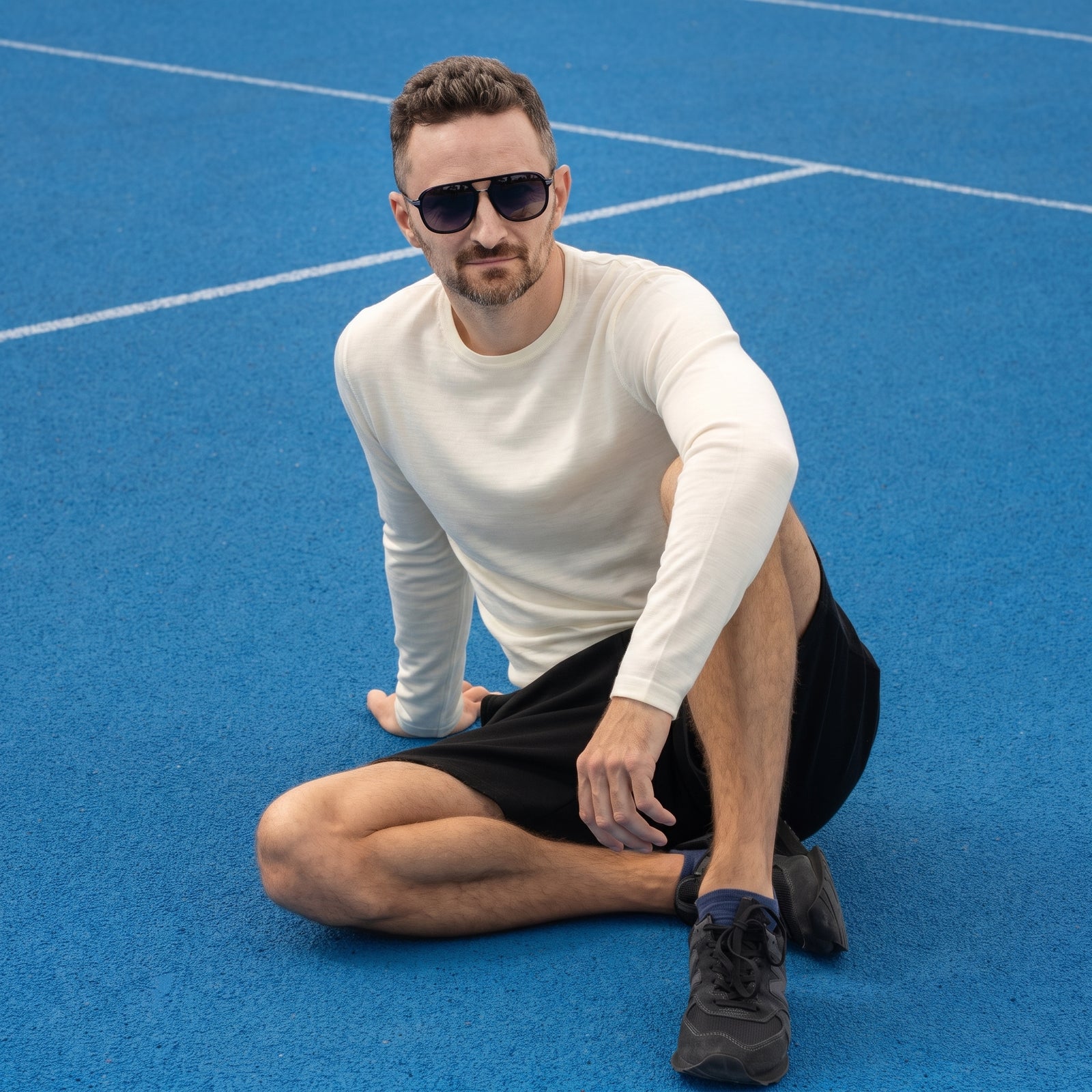 A man wearing a long-sleeved cream shirt and black shorts, sitting casually on a bright blue sports court next to a dark wire fence.