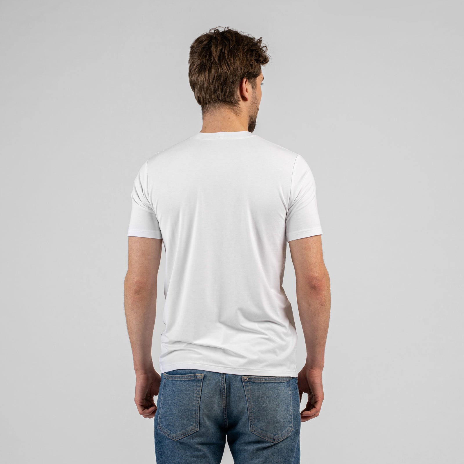 A studio portrait of a man looking directly at the camera with his hands resting behind his back. He is wearing a classic white short-sleeved t-shirt and blue jeans against a light grey background.