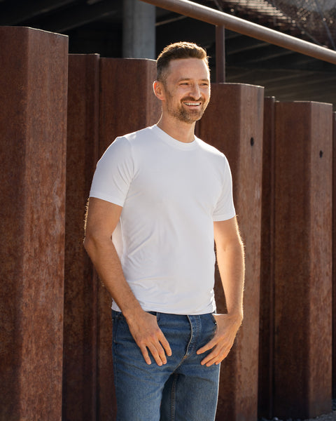 A smiling man with short brown hair and a beard stands outdoors in front of tall, vertical rusted steel beams. He is wearing a plain white short-sleeved t-shirt and blue jeans, with his hands resting casually near his front pockets, looking off to the right side of the frame.