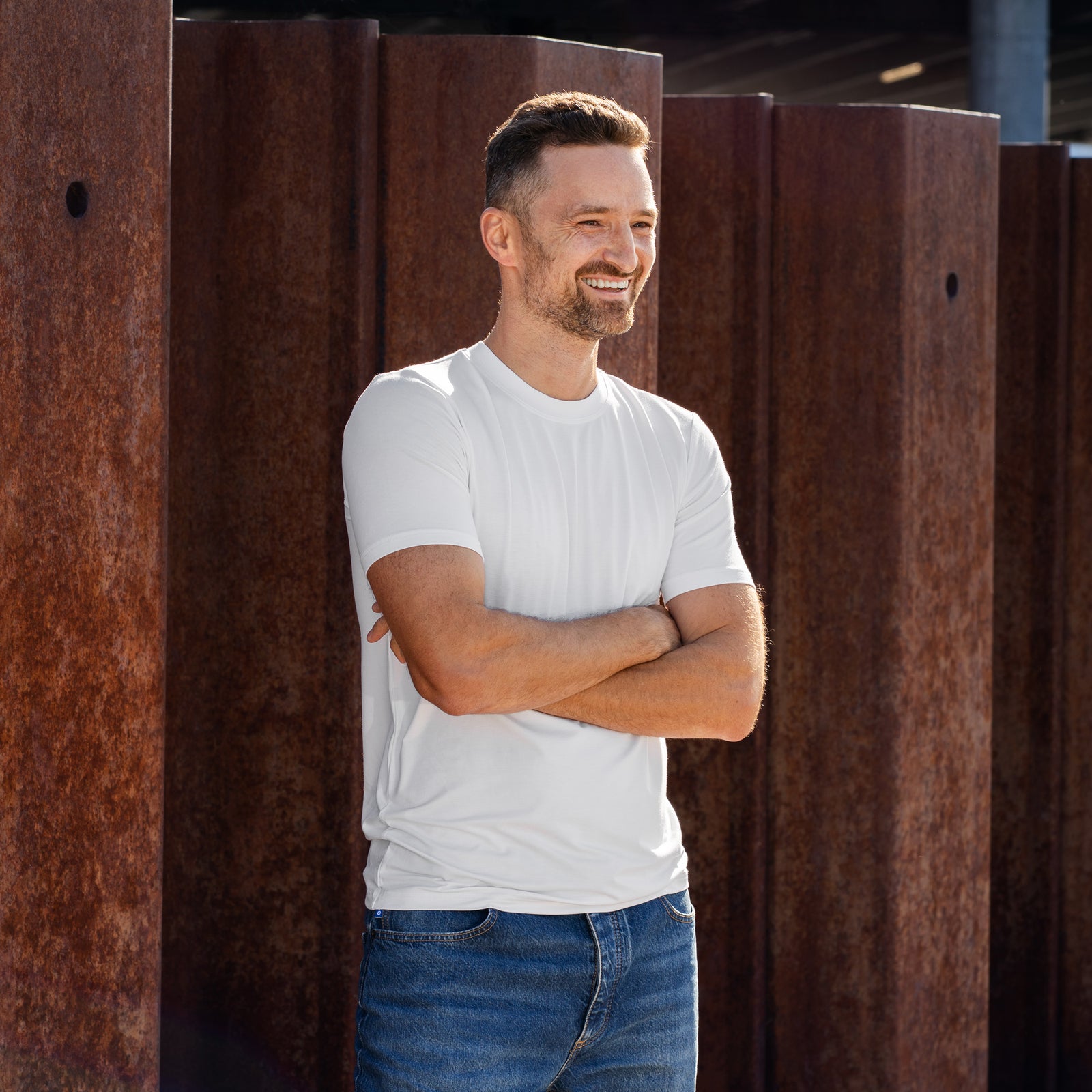 A man stands outdoors against a backdrop of rusted steel beams and a modern concrete overpass. He is wearing a plain white short-sleeved t-shirt and blue jeans. His right hand rests on his hip, and his left hand is tucked into his front pocket as he looks off into the distance.